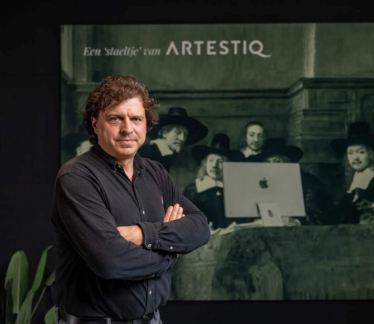 Man kijkt in camera met armen over lkaar voor een schilderij