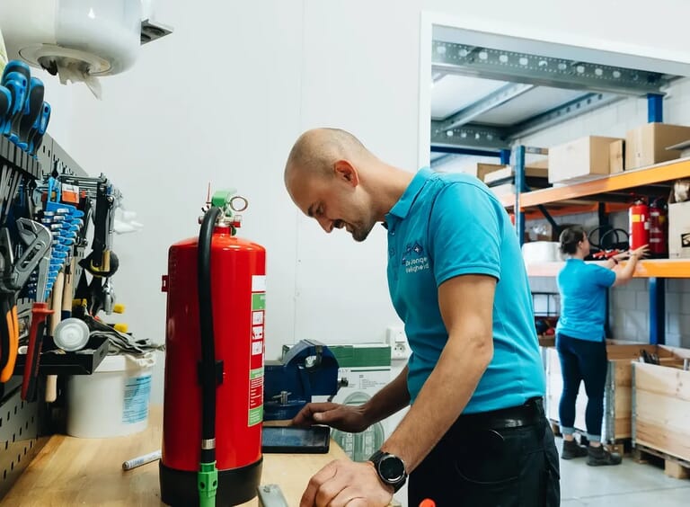 Een man die op een tablet kijkt en een brandblusser naast zich heeft staan en in de verte zie je een vrouw bij een magazijnstelling