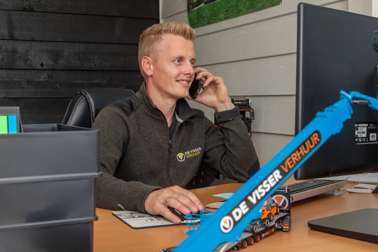 Man met De Visser Verhuur-kleding belt vanuit een kantoorruimte, met een computerscherm en schaalmodel van een hoogwerker op het bureau.