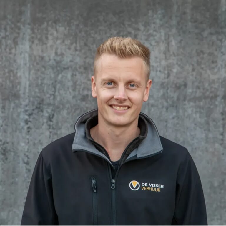 Man in jas met logo van De Visser Verhuur, staand voor een grijze betonnen achtergrond, lachend in de camera.