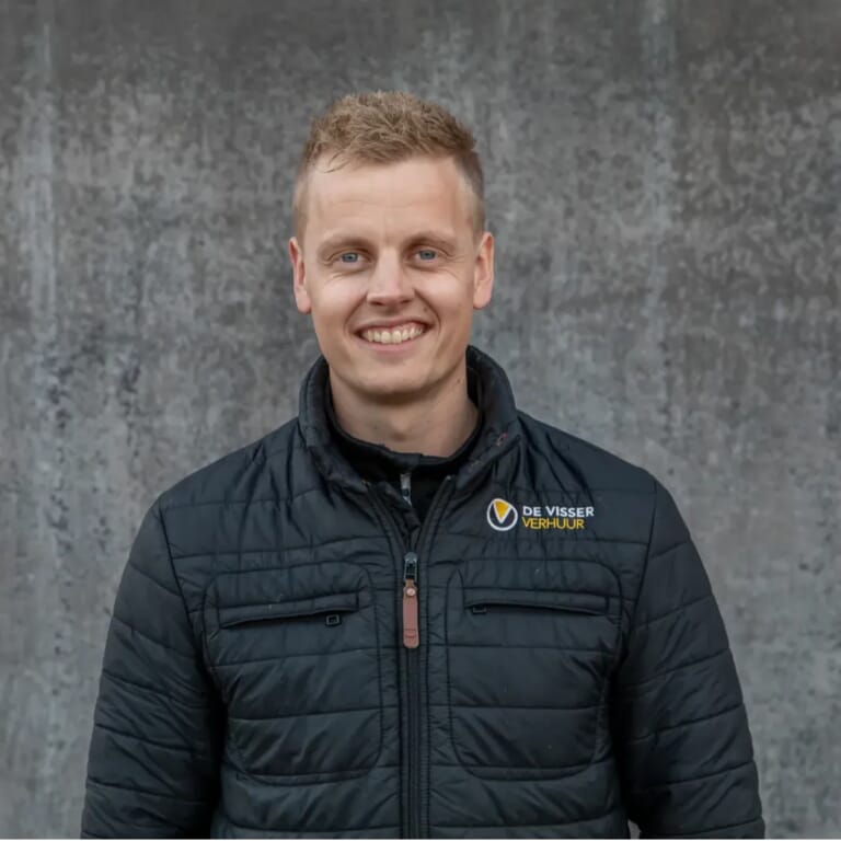 Man in gewatteerde jas met De Visser Verhuur-logo, lachend voor een grijze betonnen muur.