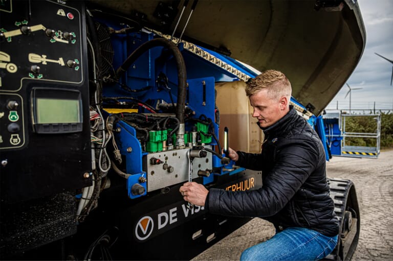 Monteur voert onderhoud uit aan een hoogwerker op een werklocatie in de buitenlucht, met gereedschap en inspectielamp in de hand, terwijl de