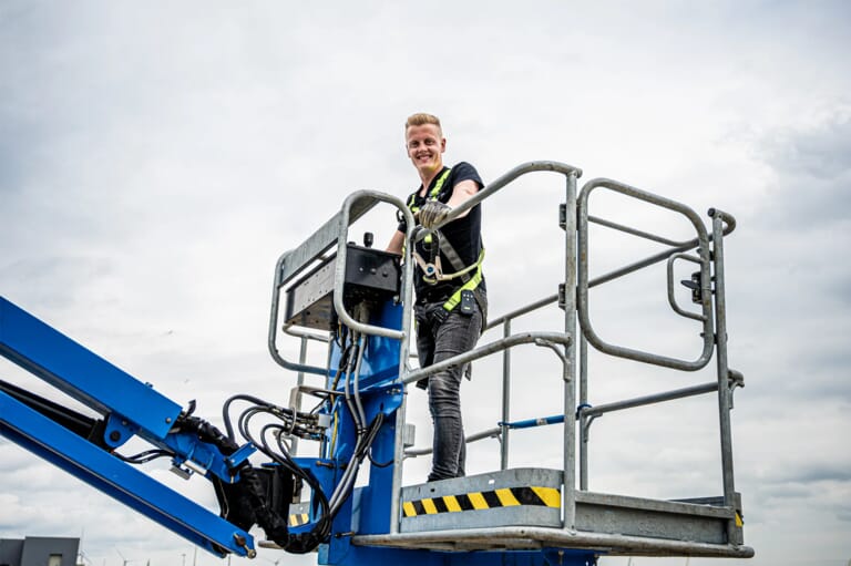 Monteur staat met veiligheidsharnas in de werkbak van een blauwe hoogwerker op hoogte, klaar voor inspectie of onderhoudswerkzaamheden in de