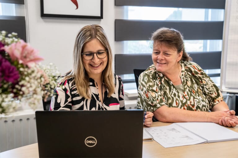Twee vrouwen achter een laptop met een opengeslagen boek