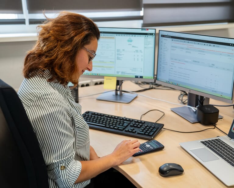 Vrouw aan een bureau met beeldschermen en tikt met haar vinger op een calculator