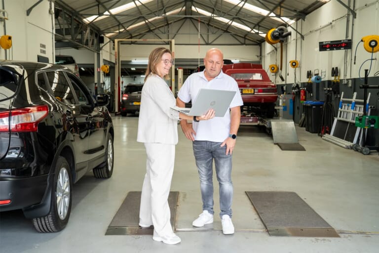Een vrouw en een man in een garage. De vrouw houdt een laptop vast en de man kijkt mee