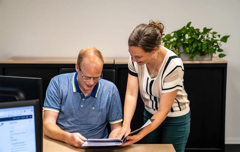 Een man zit op een stoel aan een bureau, met daarnaast een vrouw die staat met een document en dit laat zien aan de man op de stoel