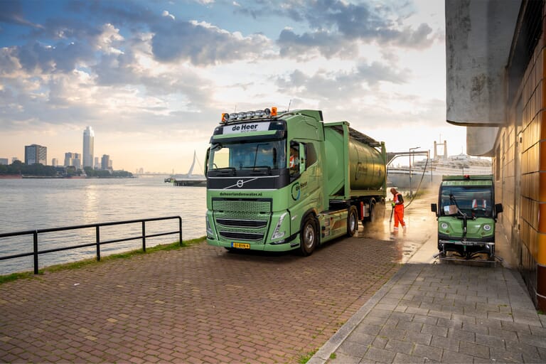 Een vrachtwagen met daarnaast en kleine auto en iemand die de vrachtwagen schoon spuit