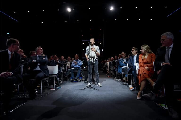 Man achter een microfoon in een grote zaal met mensen