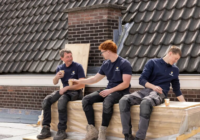 Drie mannen in werkuniforms drinken koffie op een balk op een dak