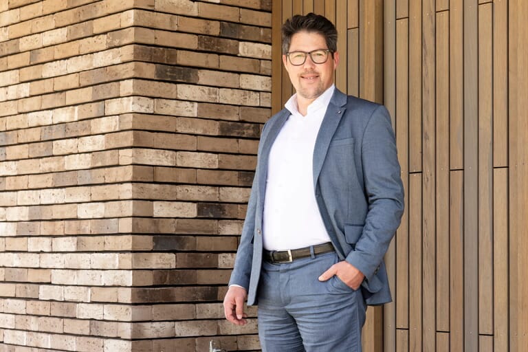 Man in blauw pak bij bakstenen en houten muur.