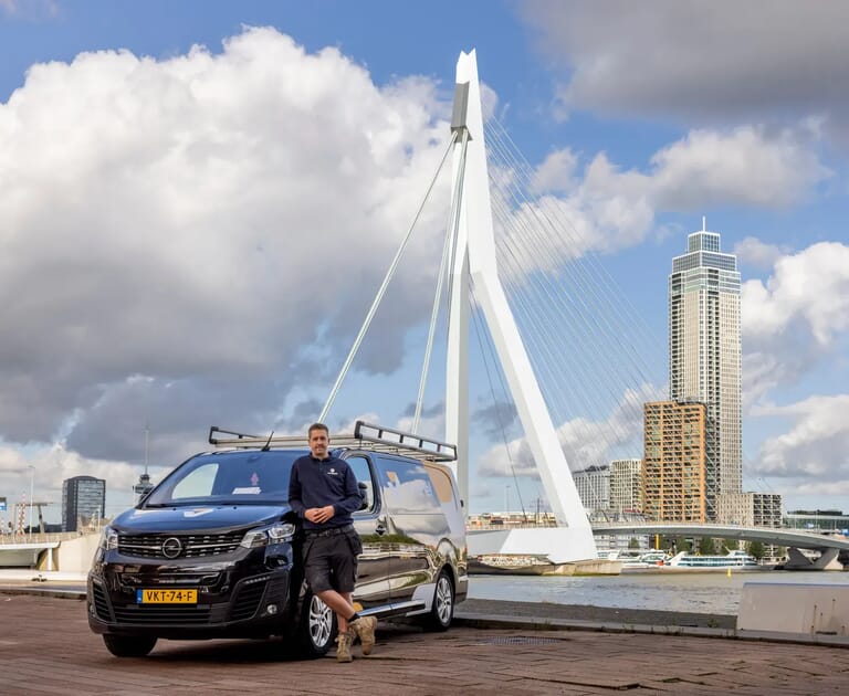 Man leunt tegen een zwarte bestelwagen bij een brug met een moderne stad op de achtergrond.