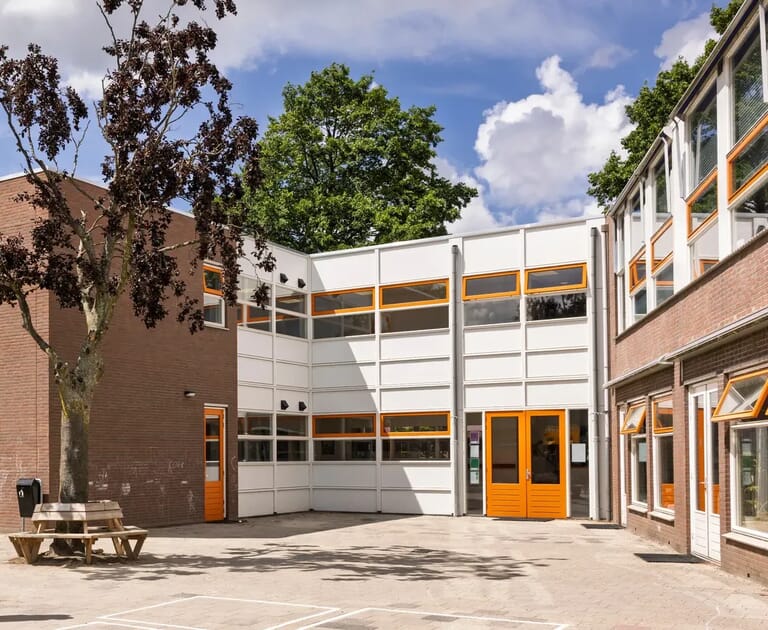 Schoolplein met een bakstenen gebouw en oranje deuren onder een blauwe hemel.