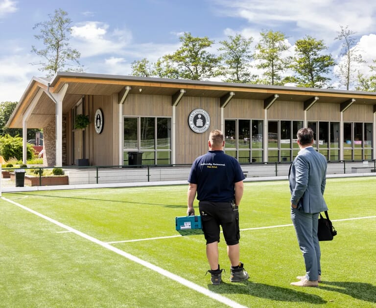 Twee mannen bekijken een modern gebouw naast een voetbalveld.