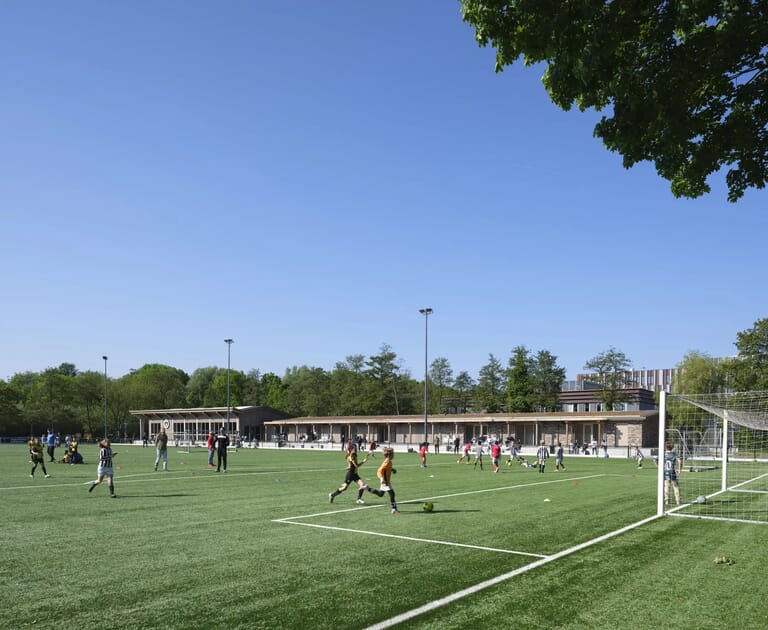 Kinderen spelen voetbal op een kunstgrasveld bij een clubhuis.