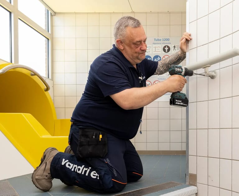 Man repareert leuning met accuboormachine bij gele glijbaan.