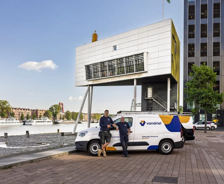 Twee mannen bij een vandriel bestelwagen voor een modern gebouw aan het water.