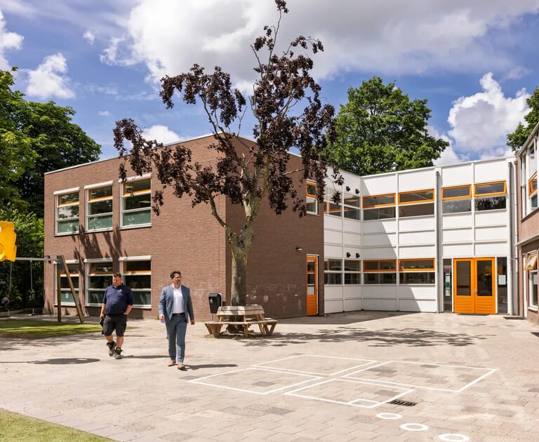 Twee mannen lopen op een schoolplein met een boom en bakstenen gebouw.