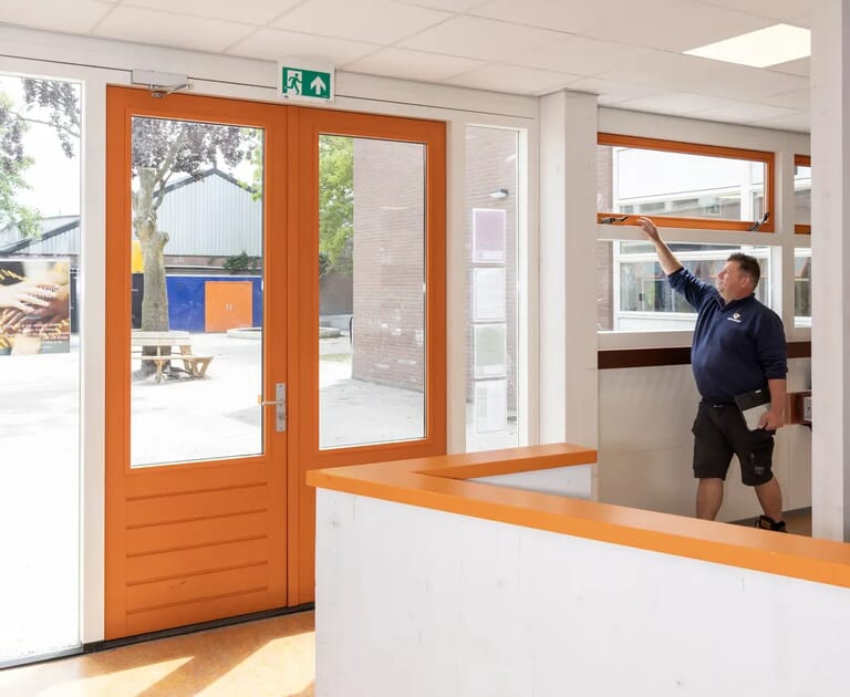 Man opent raam in een gebouw met oranje deuren.