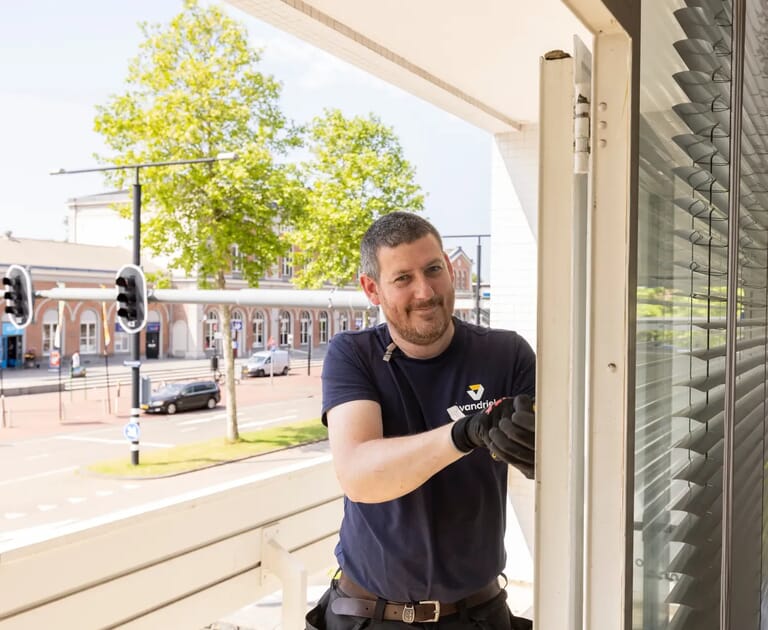 Man repareert raam op balkon met stadsverkeer op de achtergrond.