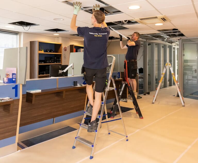Twee mannen werken aan het plafond van een kantoor met ladders.