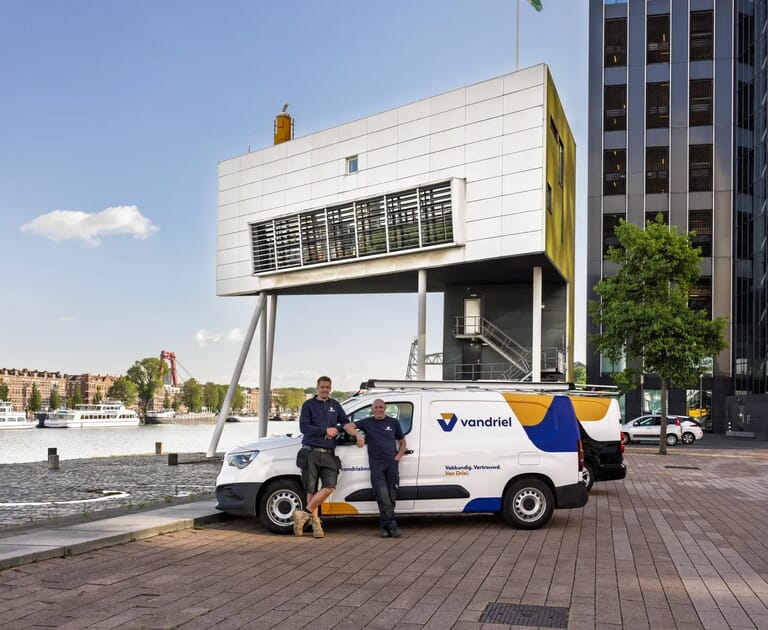 Twee mannen leunen tegen een bedrijfswagen voor een modern gebouw aan het water.