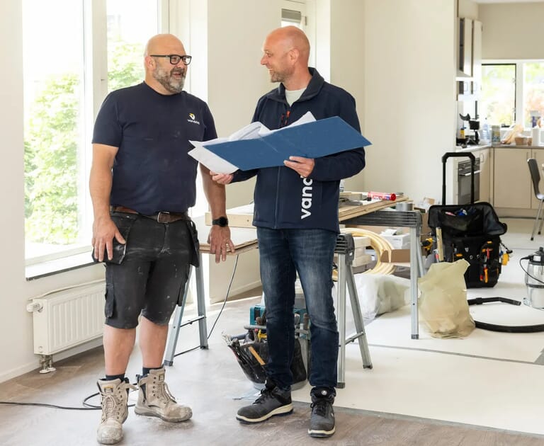 Twee mannen overleggen in een werkplaats met bouwmateriaal.