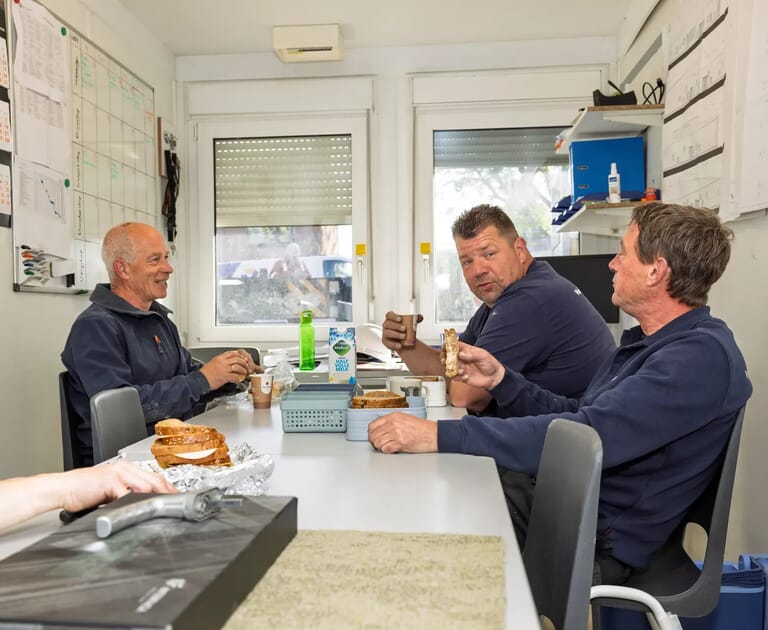 Drie mannen lunchen en praten in een kantoorruimte.